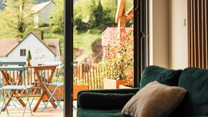 A room with a green sofa, a wooden table with two wine glasses, and a view of houses and trees outside. - Vrbo Property (Bernardvillé)