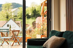 Une pièce avec un canapé vert, une table en bois sur laquelle reposent deux verres à vin, et une vue sur les maisons et les arbres à l’extérieur.