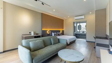 A modern hotel room with a sofa, a round table, a bed, a desk, and a bathroom with a mirror.