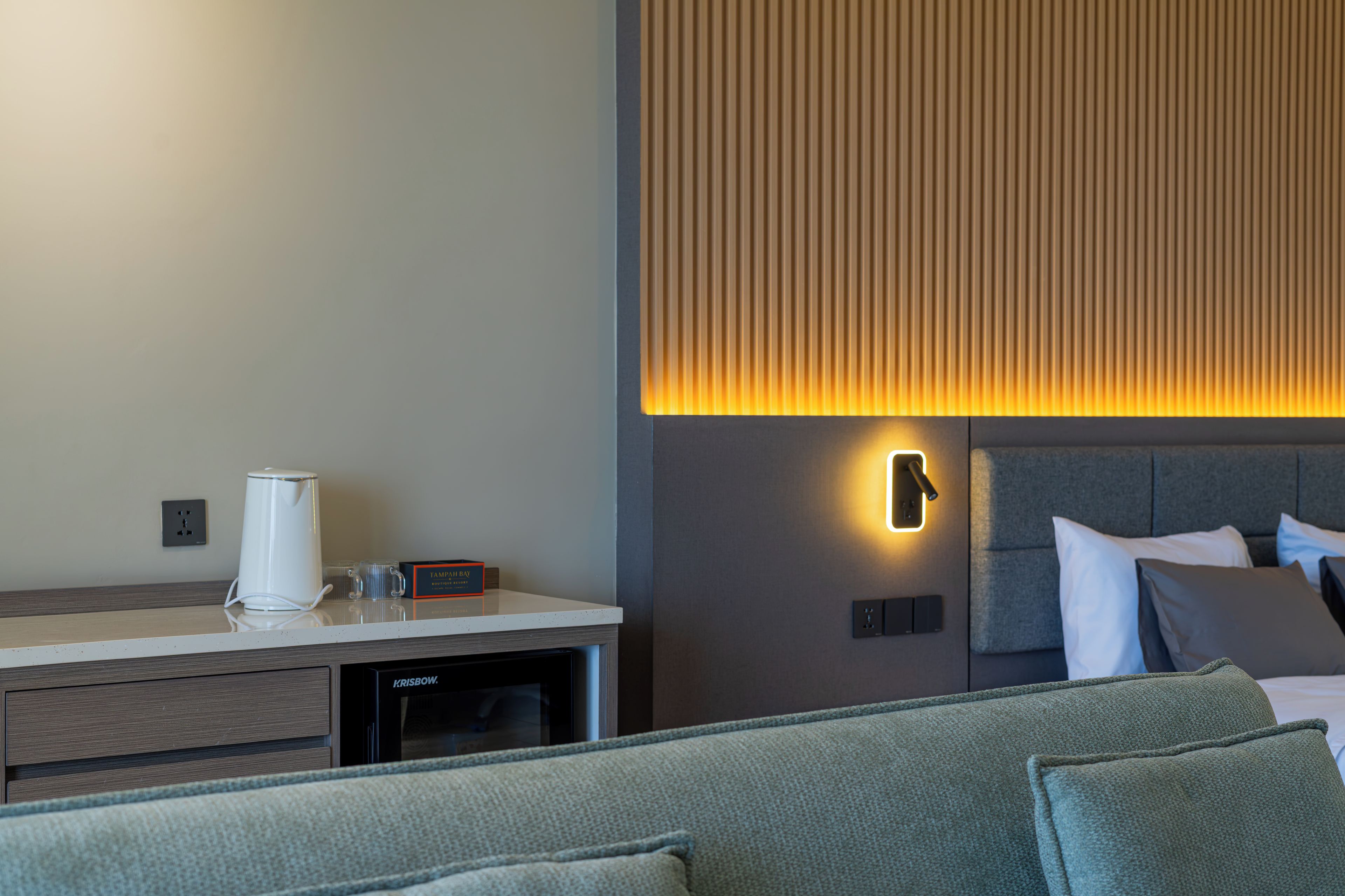 A modern hotel room with a bed, a bedside table with a coffee maker, and a wall-mounted light.