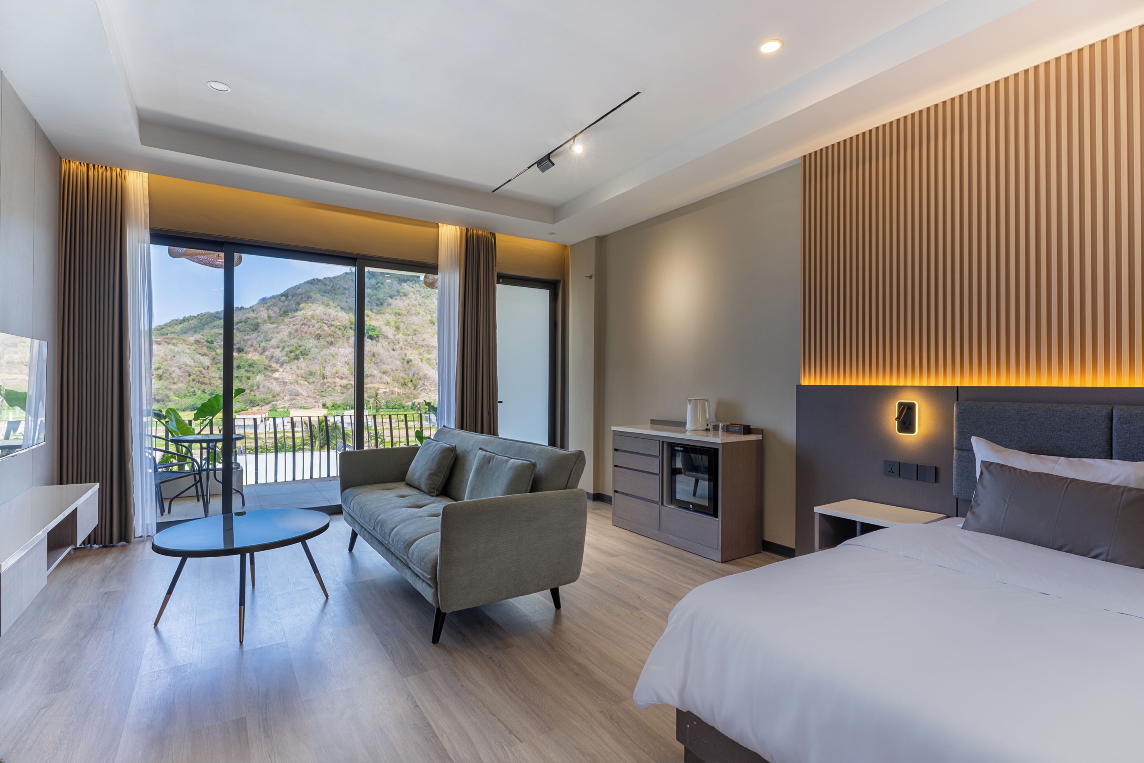 Una habitación de hotel moderna con balcón, sofá, mesa de comedor, cama y una pequeña kitchenette.