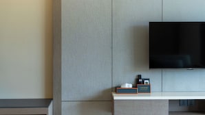A modern hotel room with a flat-screen TV mounted on a grey wall, a small table with a remote control and a tissue box, and a wooden nightstand. - Tampah Bay Boutique Resort (Kuta)