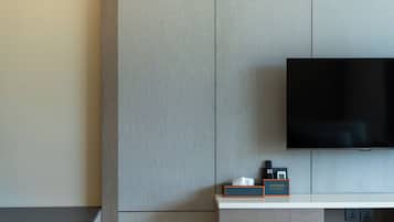 Moderni hotellihuone, jossa on seinälle asennettu litteänäyttöinen televisio, pieni pöytä, jossa on kaukosäädin ja paperinenäliöpakkaus, sekä puinen yöpöytä.