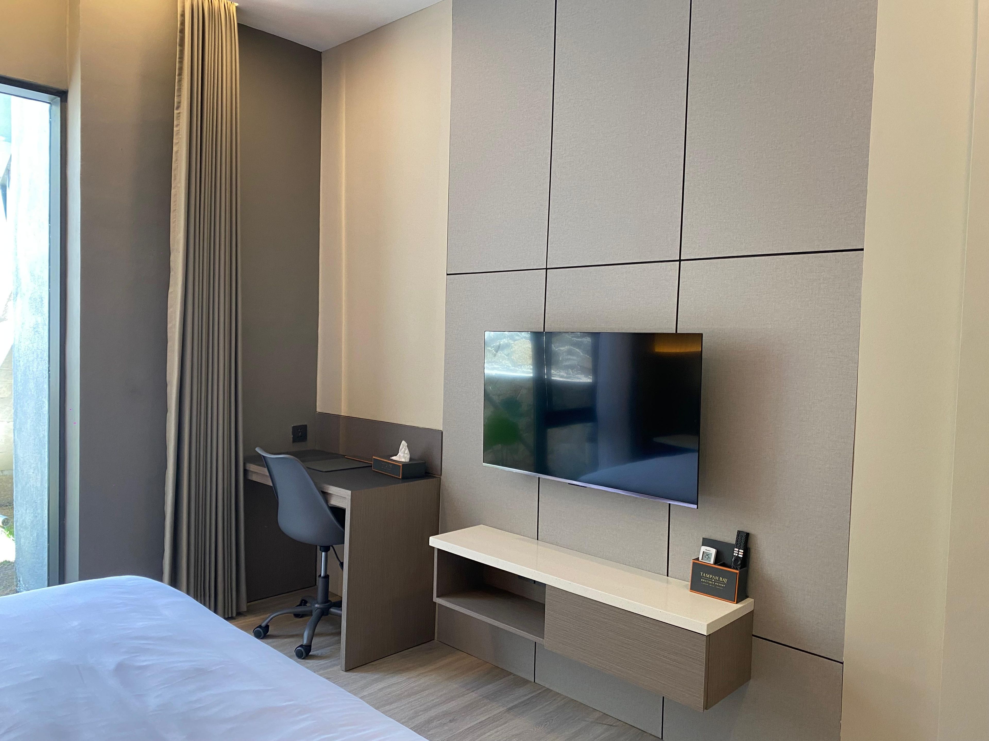 Una habitació d'hotel moderna amb un llit, escriptori, cadira, televisió penjada a la paret i finestra amb cortines.