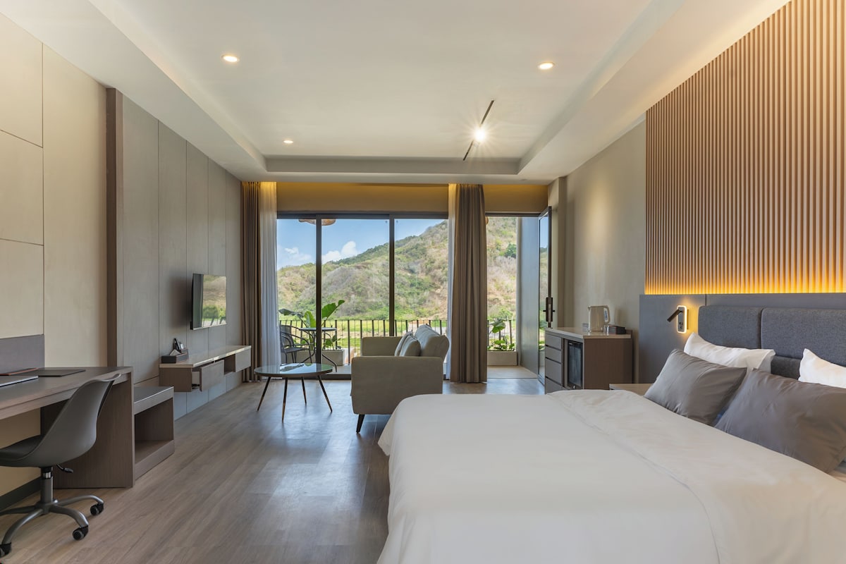 A hotel room with a large bed, a desk, a chair, a television, and a view of a mountainous landscape.