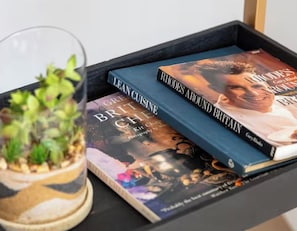 A glass terrarium with plants, two books titled 'Great British Cuisine' and 'Rhodes Around Britain,' and a black tray. - J-Fitzrovia Skyhome | Spacious Central London 2BR (London)