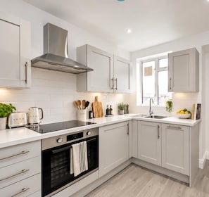 A modern kitchen with white cabinetry, a stainless steel range hood, a black oven, and a window providing natural light. - J-Fitzrovia Skyhome | Spacious Central London 2BR (London)