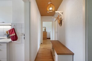 A hallway with a wooden floor, white walls, a kitchen area with a red towel, and a hanging light fixture.