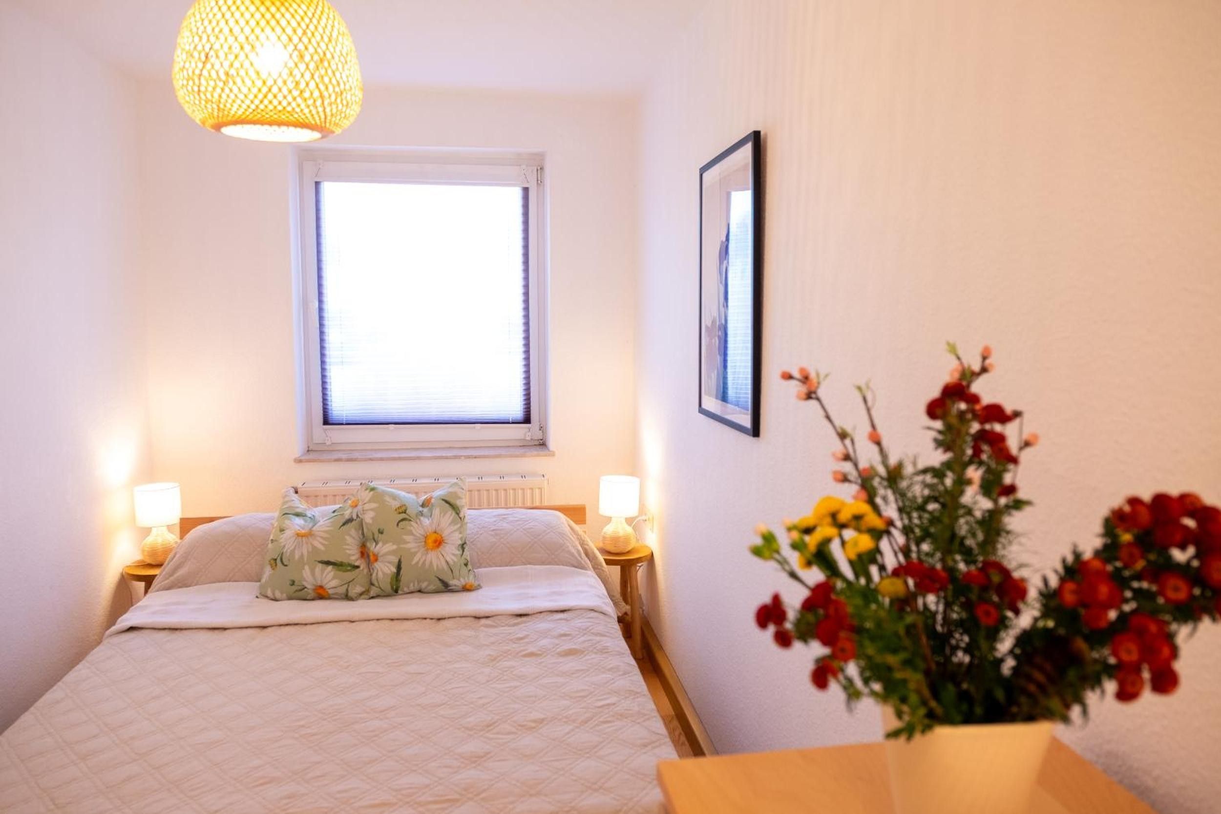 Ein Schlafzimmer mit einem Bett, zwei Nachttischlampen, einem Fenster, einem gerahmten Bild und einer Vase mit Blumen.