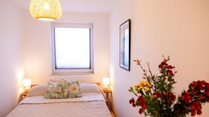 Ein Schlafzimmer mit einem Bett, zwei Nachttischlampen, einem Fenster, einem gerahmten Bild und einer Vase mit Blumen.