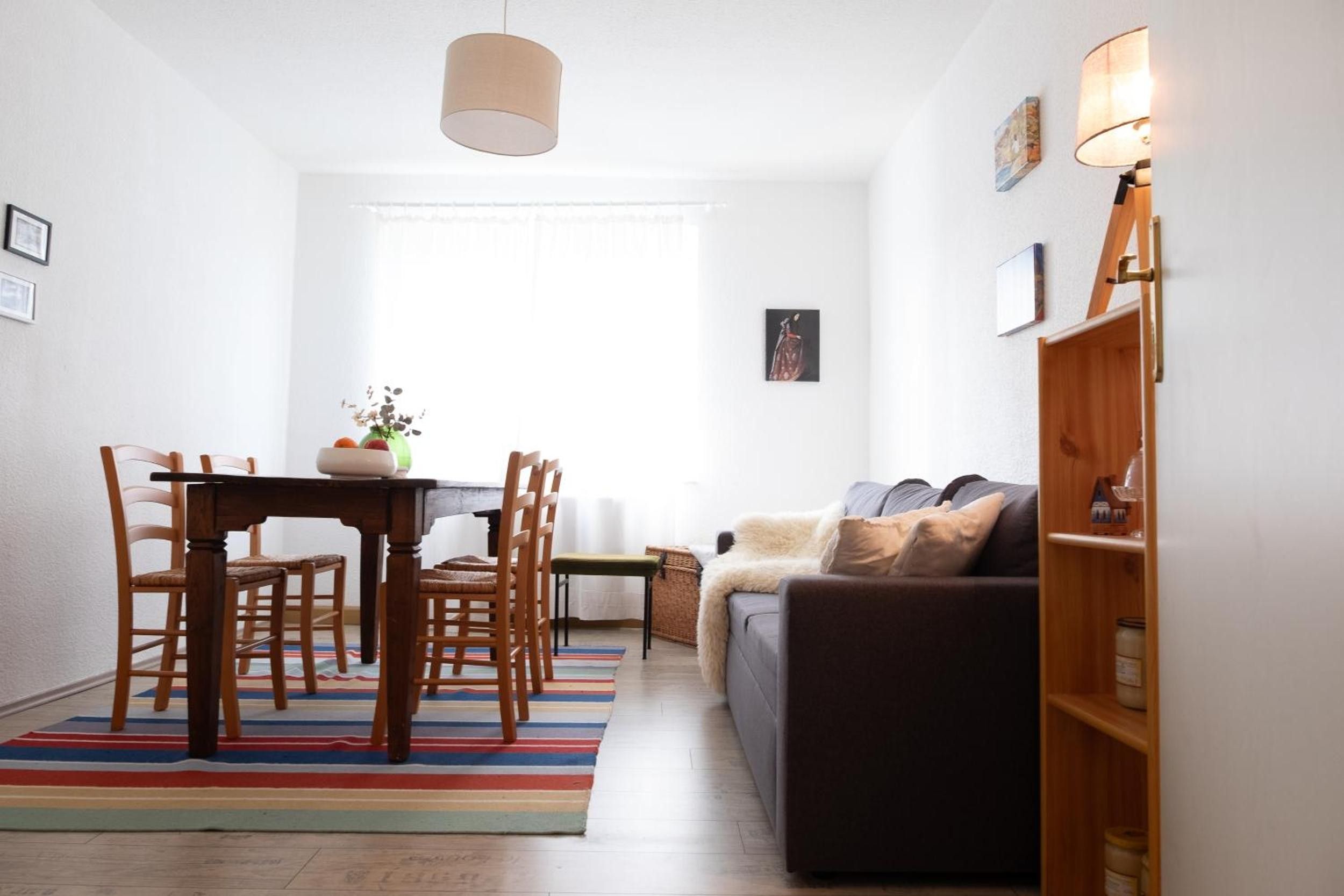 Ein modernes Wohnzimmer mit einem dunklen Sofa, einem Holzesstisch und Stühlen. Ein farbenfroher Streifen Teppich, ein kleiner Beistelltisch und ein Fenster mit weißen Vorhängen sind ebenfalls vorhanden.