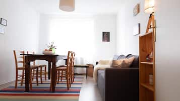 Ein modernes Wohnzimmer mit einem dunklen Sofa, einem Holzesstisch und Stühlen. Ein farbenfroher Streifen Teppich, ein kleiner Beistelltisch und ein Fenster mit weißen Vorhängen sind ebenfalls vorhanden.