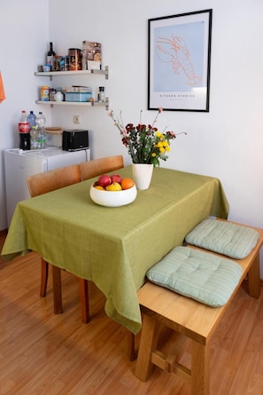A dining area with a table covered by a green tablecloth, a bowl of fruit, and a potted plant with yellow flowers.