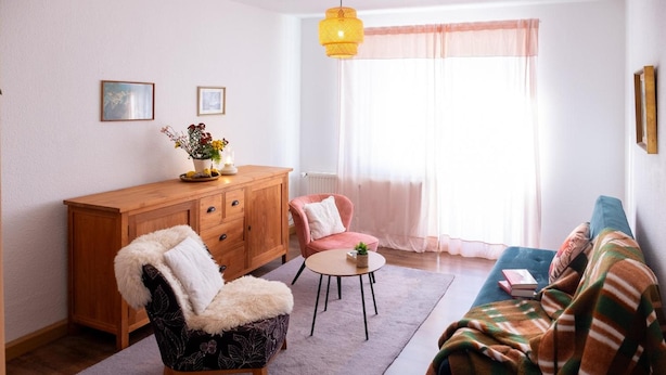 Ein Wohnzimmer mit einem Holzkabinett, einem Sessel mit Felldecke, einem runden Couchtisch, einer Couch und einem Fenster mit transparenten Vorhängen.