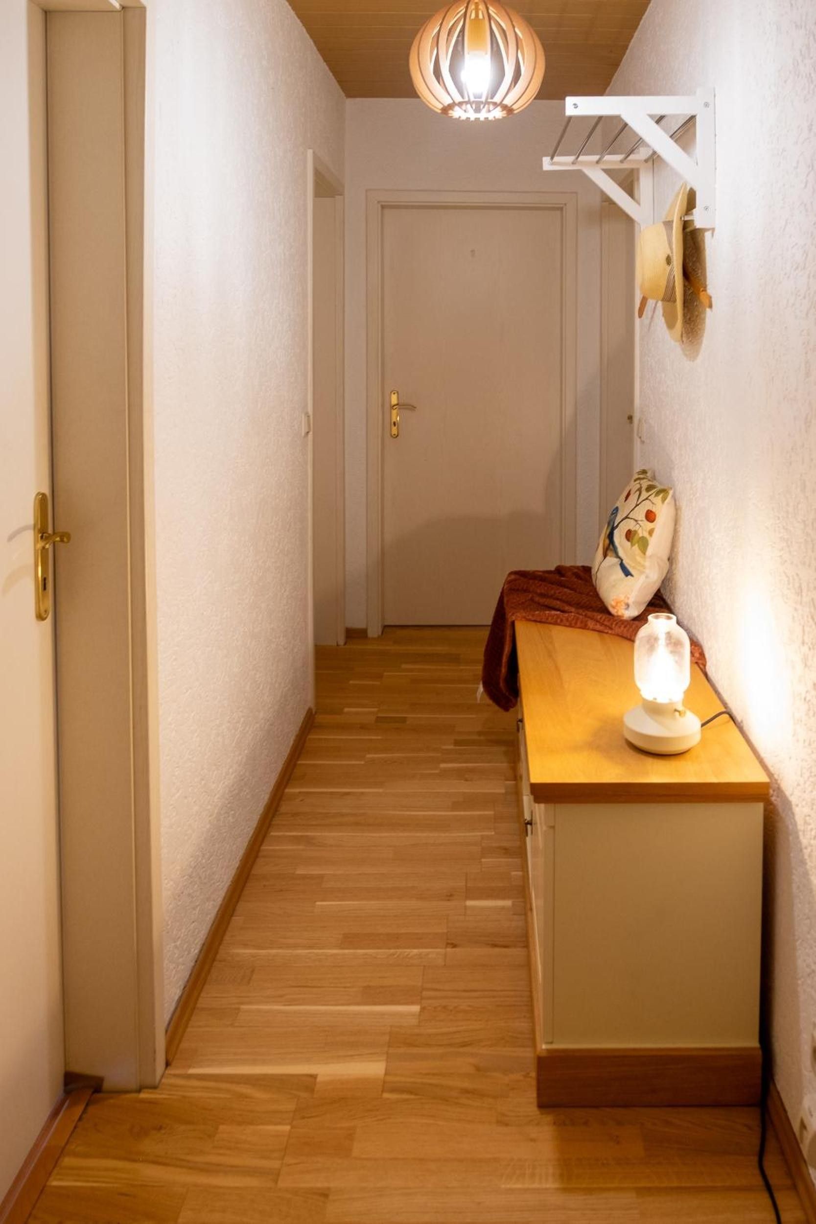 Ein schmaler Flur mit Holzboden, einer weißen Tür am Ende, einem kleinen Tisch mit Lampe und einem Wandventilator.