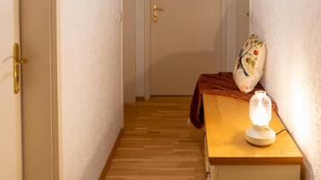 Ein schmaler Flur mit Holzboden, einer weißen Tür am Ende, einem kleinen Tisch mit Lampe und einem Wandventilator.