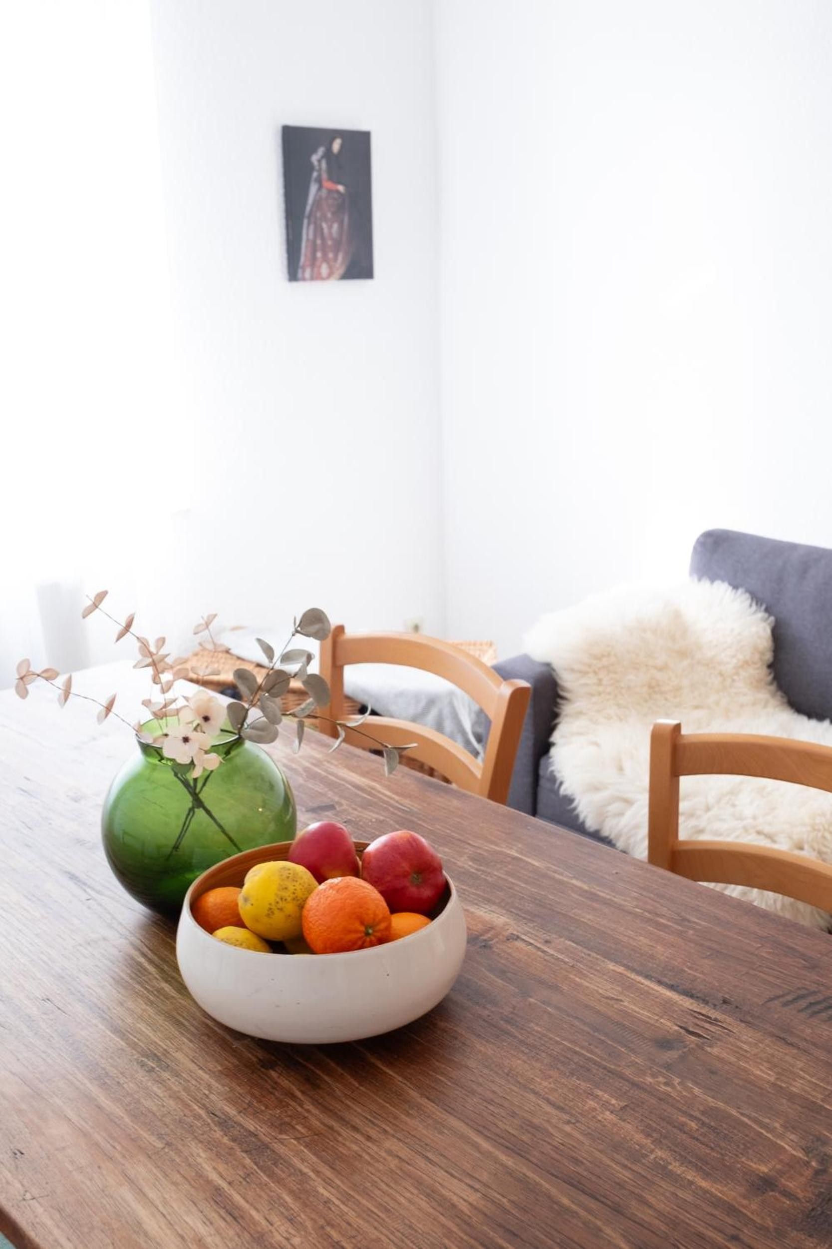 Ein Esstisch mit Obstschale und einer grünen Vase mit Zweigen.
