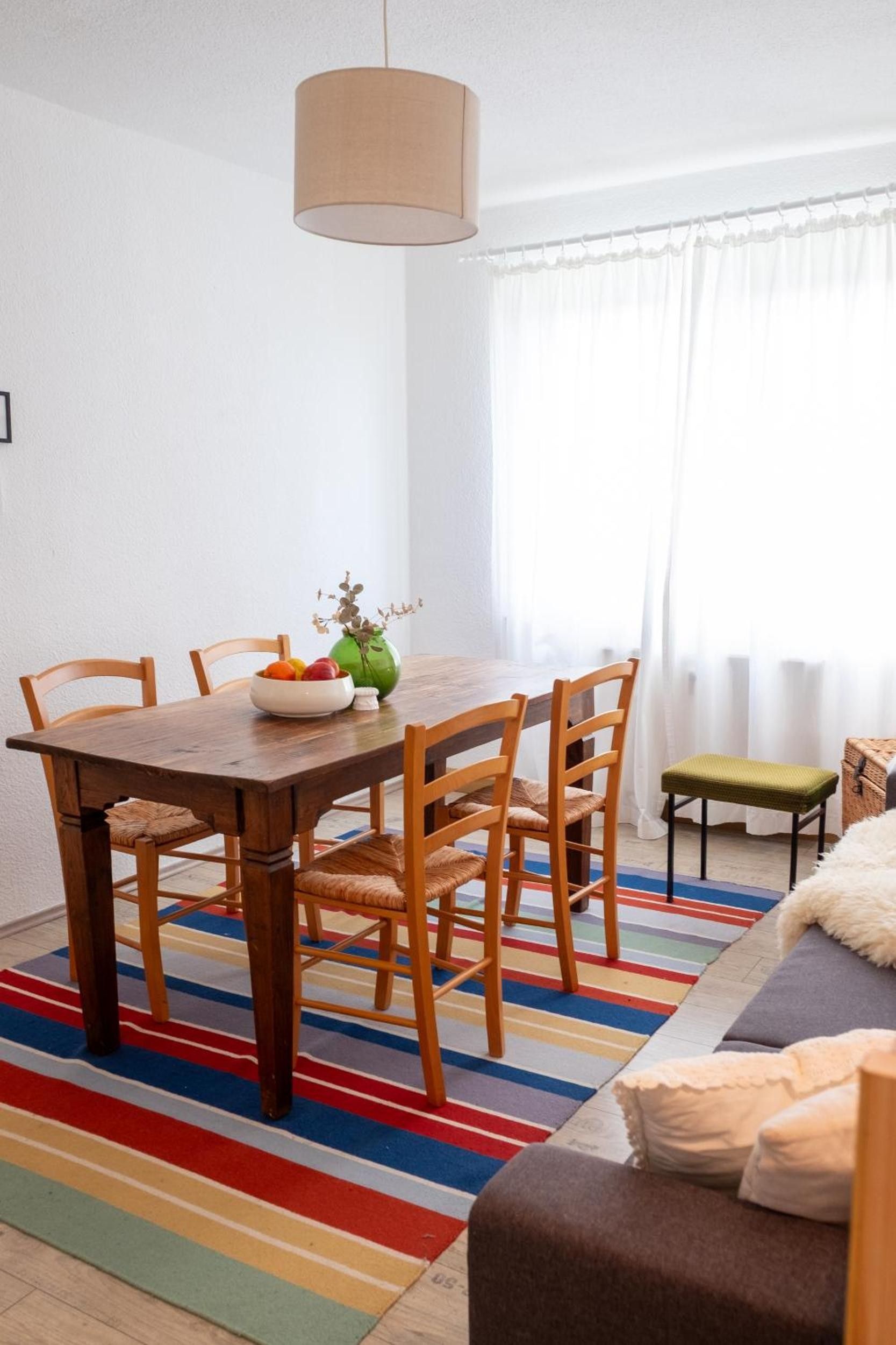 Ein Essbereich mit einem Holztisch, Stühlen, einer Blumenvase und einem farbenfrohen Streifen Teppich.