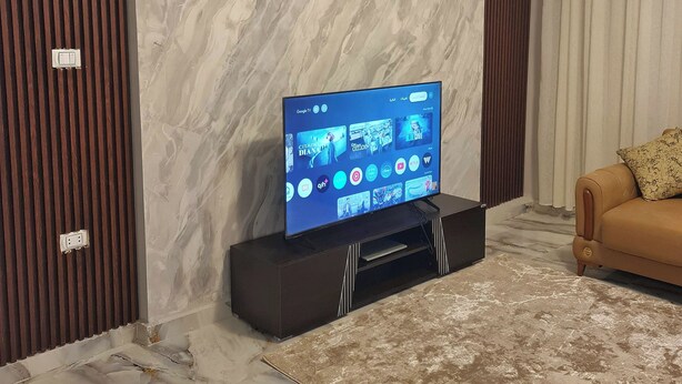 Een woonkamer met een flatscreen televisie op een standaard, een bruine leren bank en een vloer met een marmerpatroon.