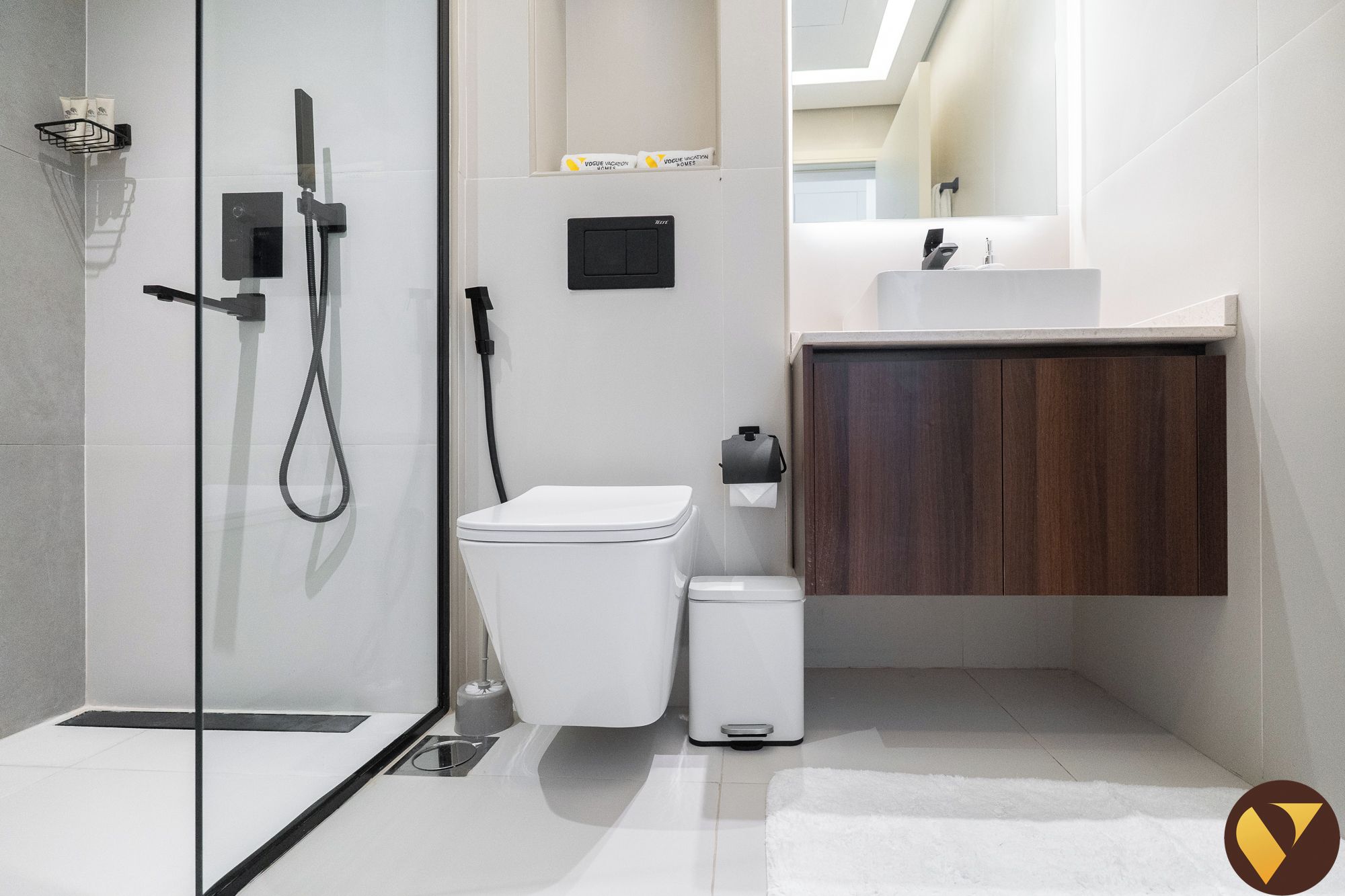 Ein modernes Badezimmer mit Duschkabine aus Glas, Toilette, kleinem Mülleimer und Holzschrank über einem Waschbecken.