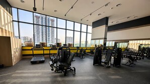 A gym with exercise equipment, large windows, and a view of high-rise buildings. - Vogue | PG One Al Furjan near Metro Station (Dubai)