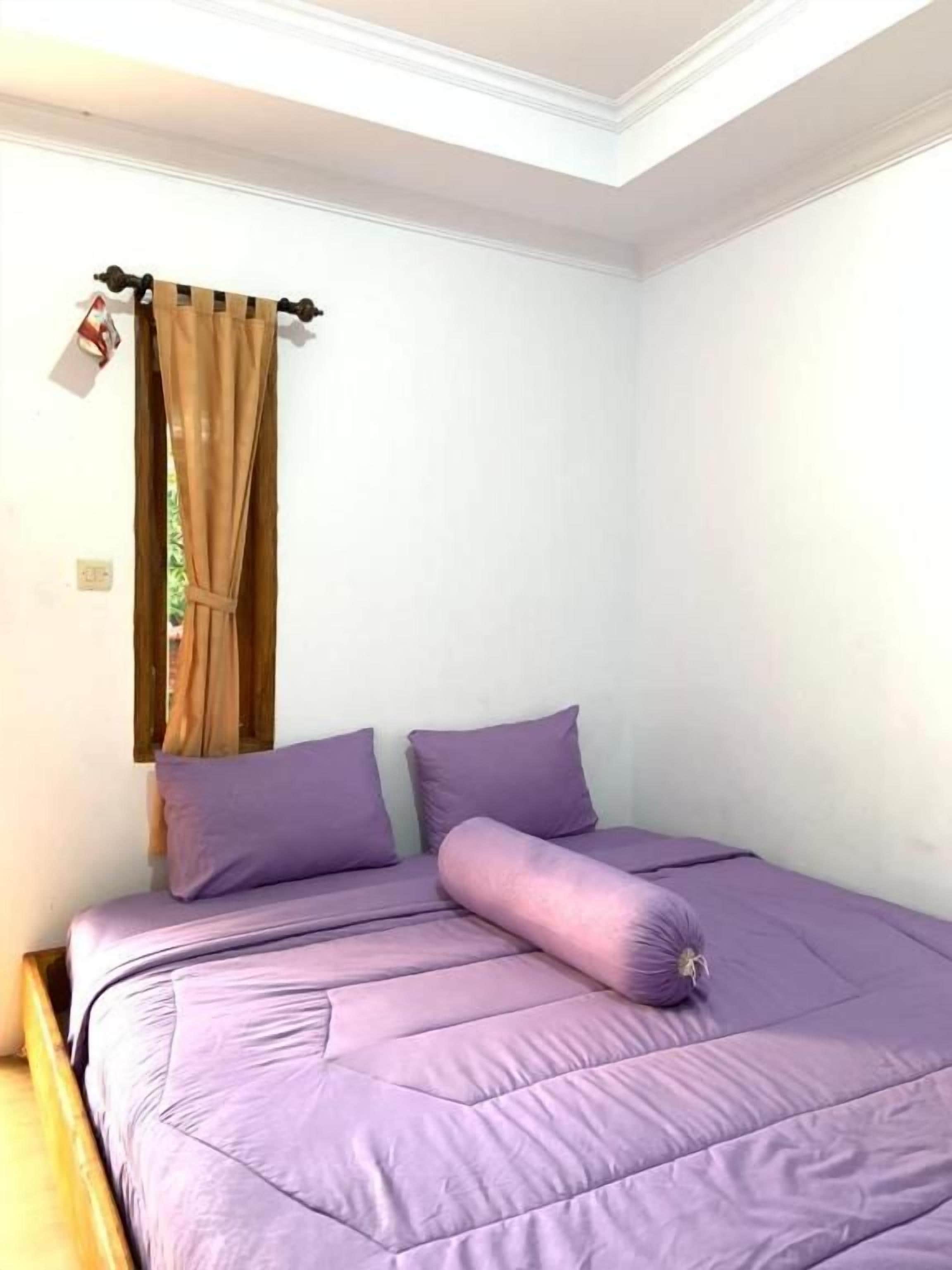 Uma cama de solteiro com colcha e almofadas roxas, uma janela com cortinas marrons e uma parede branca.