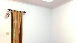 A single bed with a purple bedspread and pillows, a window with brown curtains, and a white wall. - Sokkyo Homestay Kaliurang (DI Yogyakarta)