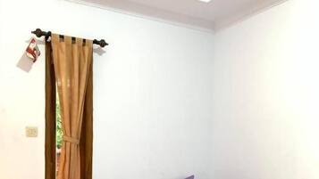 Uma cama de solteiro com colcha e almofadas roxas, uma janela com cortinas marrons e uma parede branca.