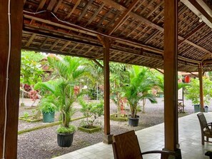 Un espacio al aire libre cubierto con pilares de madera, plantas en macetas y piso de baldosas.