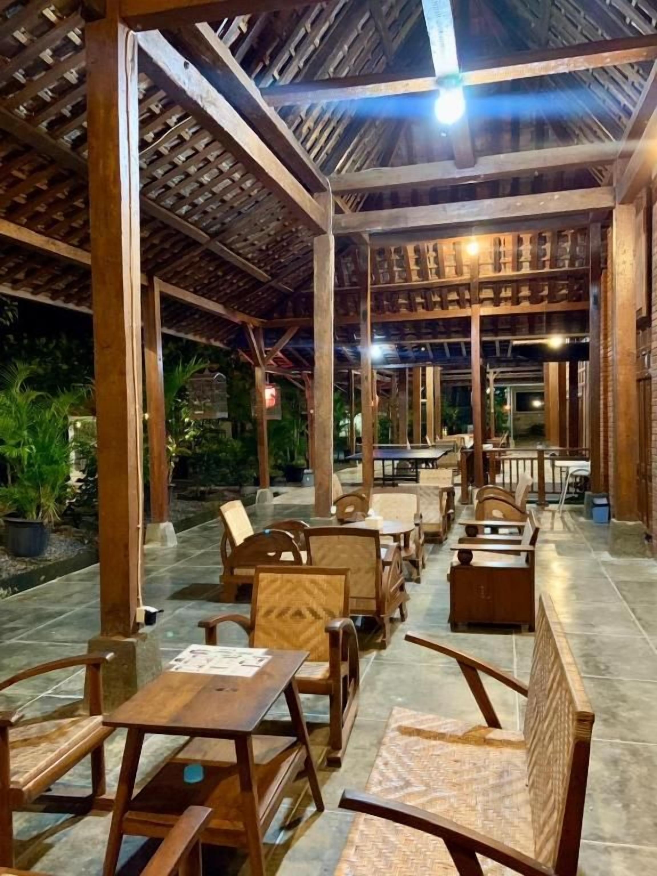 An outdoor seating area with wooden tables and chairs, a covered roof structure, and potted plants.