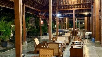 An outdoor seating area with wooden tables and chairs, a covered roof structure, and potted plants.