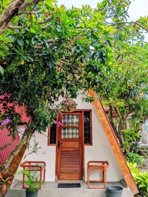 Una casa con puerta de madera, un pequeño porche con dos sillas y un árbol grande que da sombra.