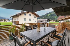 Eine Terrasse im Freien mit Tisch und Stühlen, einem Holzzaun und Blick auf ein Gebäude und eine Landschaft.