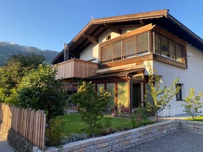 Uma casa de dois andares com varanda de madeira, um muro de pedra e um jardim com diversas plantas.