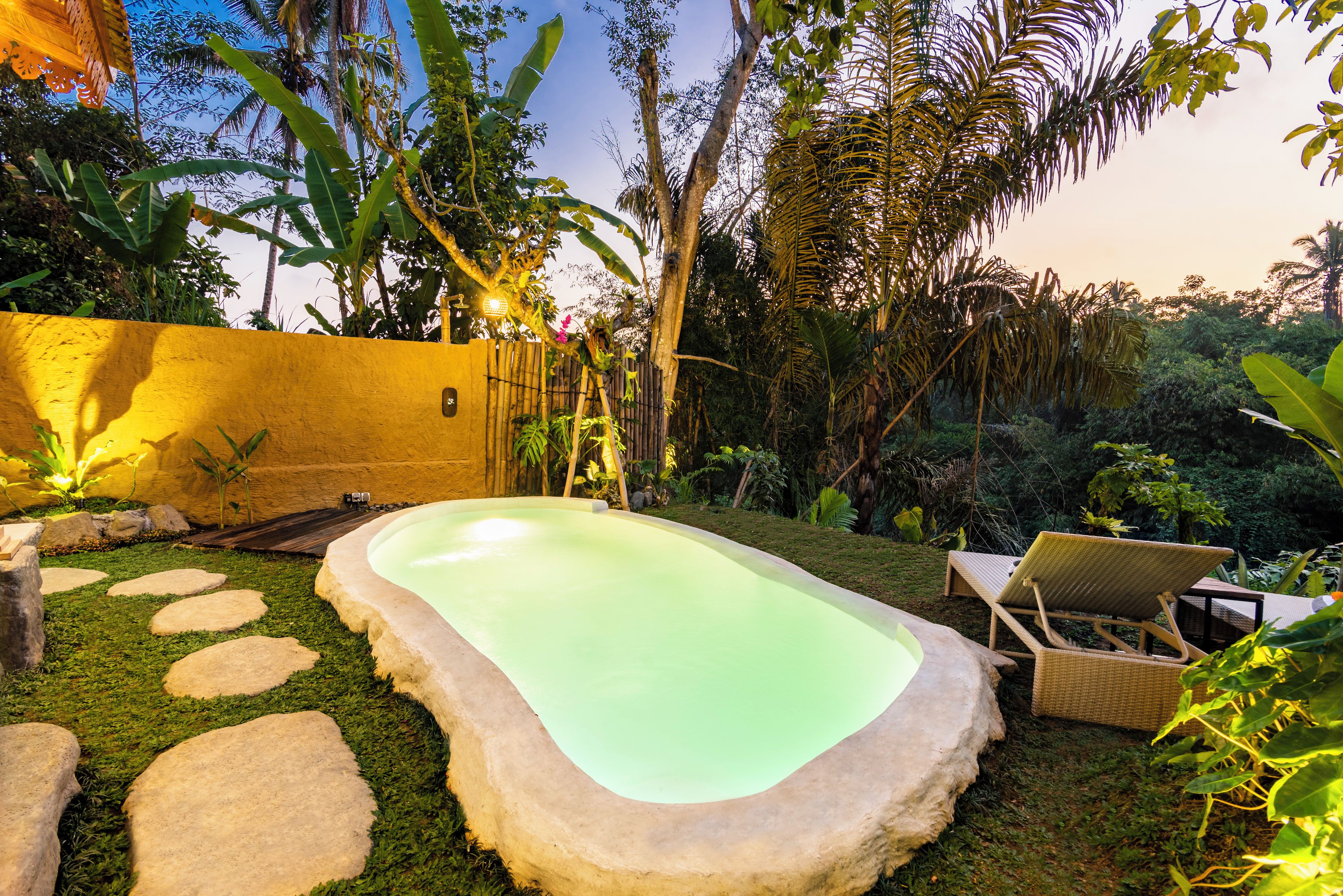 A swimming pool with a stone edge, surrounded by a grassy area with stepping stones, and a bamboo fence.