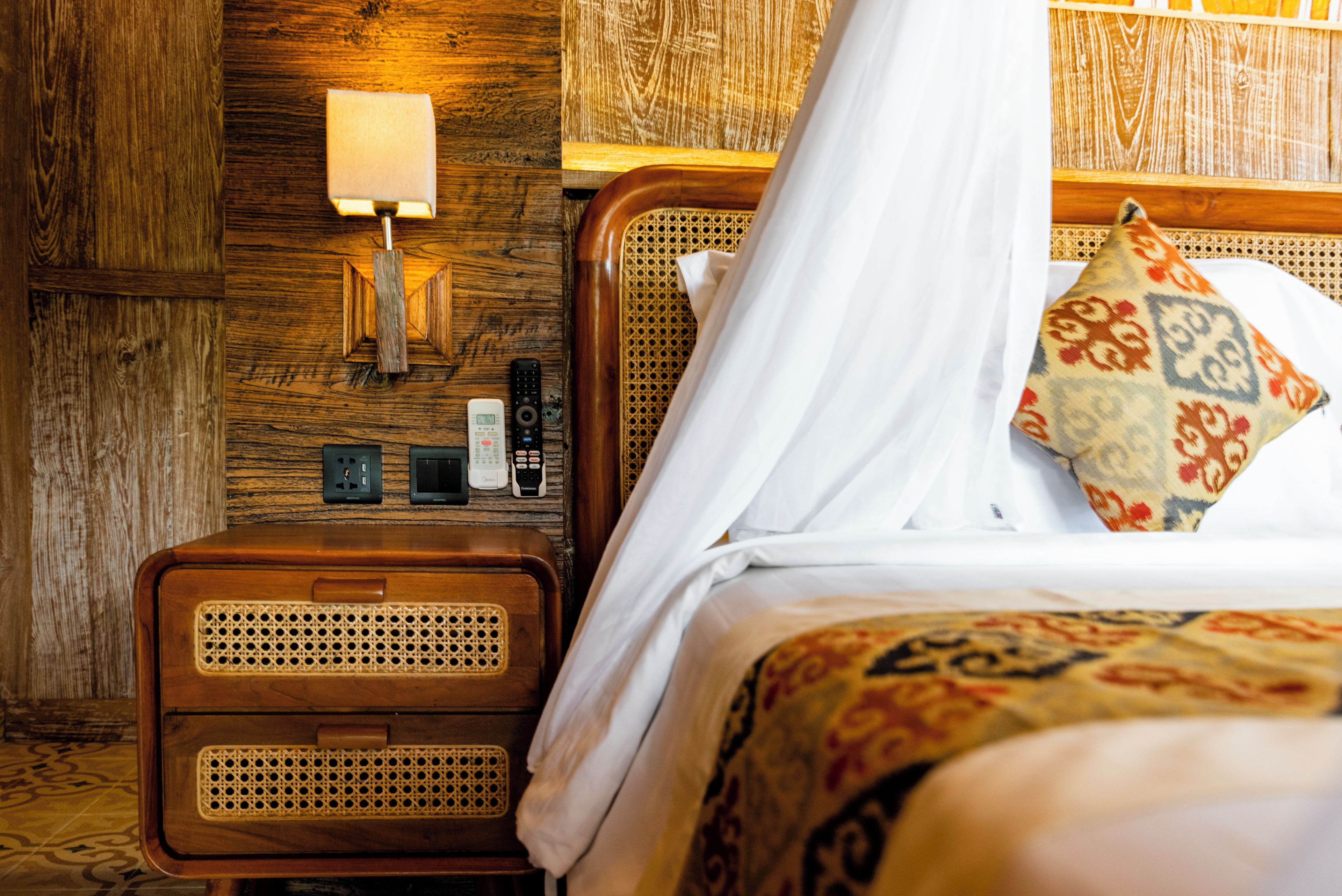 Habitación de hotel con cabecera de madera, mesita de noche con lámpara, control remoto y reloj.