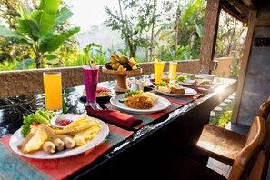 Una mesa servida con diversos platos, bebidas y una cesta de frutas, con vistas a la vegetación al fondo.