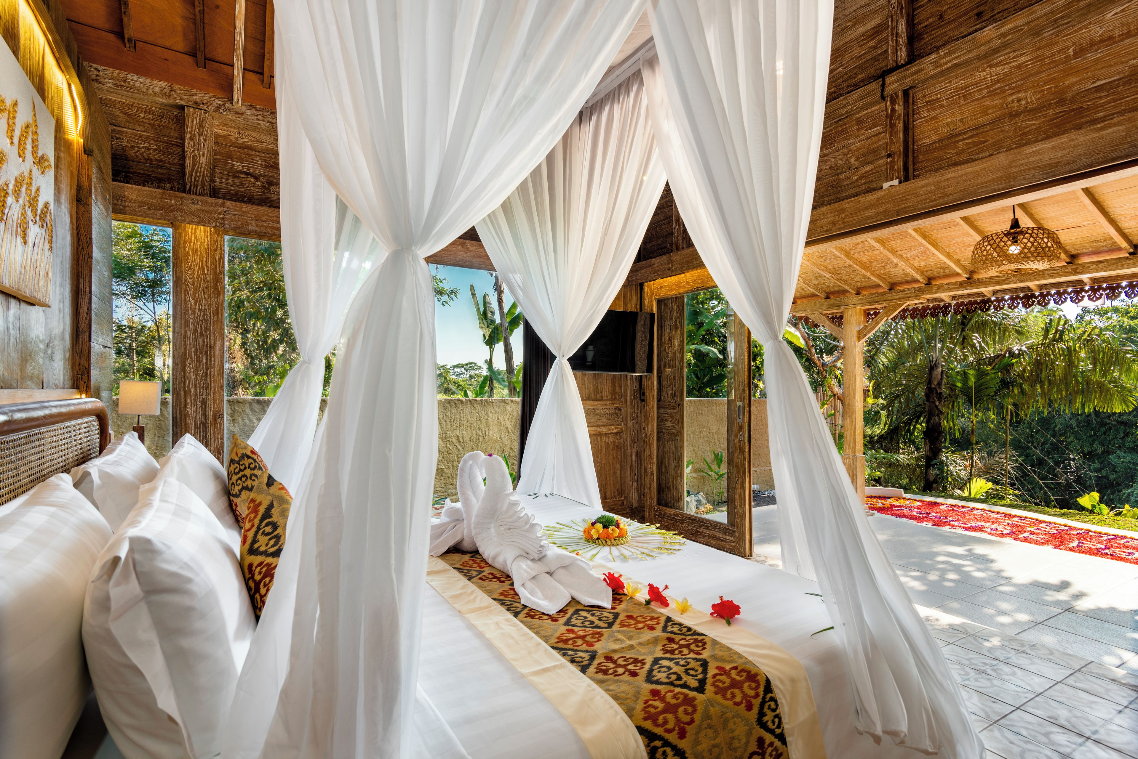 Una cama con sábanas blancas y una colcha estampada, rodeada de cortinas blancas, en una habitación con vigas de madera y vista a la vegetación.