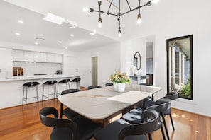 A modern kitchen with white cabinets, a marble countertop, and a dining area with a large table and chairs. - KARINYA LODGE - The Vintage Holiday Houses (POKOLBIN)