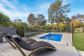 Une piscine entourée de chaises longues, d’une terrasse en bois et d’une clôture en verre.