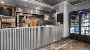 Un espace de restauration avec un présentoir de produits de boulangerie, un distributeur de boissons réfrigéré et, au fond, une cuisine en acier inoxydable.