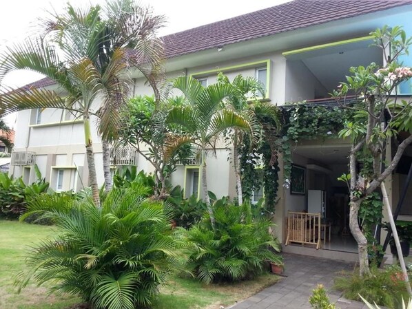 Un edificio residencial con techo de tejas, un porche cubierto y abundante vegetación, incluyendo palmeras y arbustos diversos.