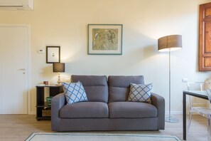 A living room with a grey sofa, two patterned throw pillows, a floor lamp, a framed artwork on the wall, a small table with a lamp, and a clear glass table to the right. - Apartments Florence - Mona Lisa Charme (Firenze)
