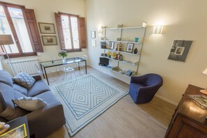 Ein Wohnzimmer mit Sofa, Sessel, Couchtisch und Teppich. Ein Bücherregal mit diversen Gegenständen und eine Heizung sind ebenfalls vorhanden.