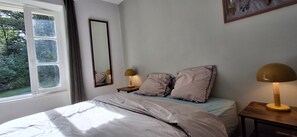A bedroom with a bed, two bedside lamps, a nightstand, a mirror, and a window with a view of greenery. - Country House 'Your Riverside Mill' with Private Terrace, Private Garden and Wi-Fi (Saint-Priest-sous-Aixe)