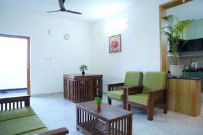 Una sala de estar con una mesa de centro de madera, dos sillones verdes, una mesita de noche de madera con una planta, un cuadro en la pared y una zona de cocina con una planta en maceta.
