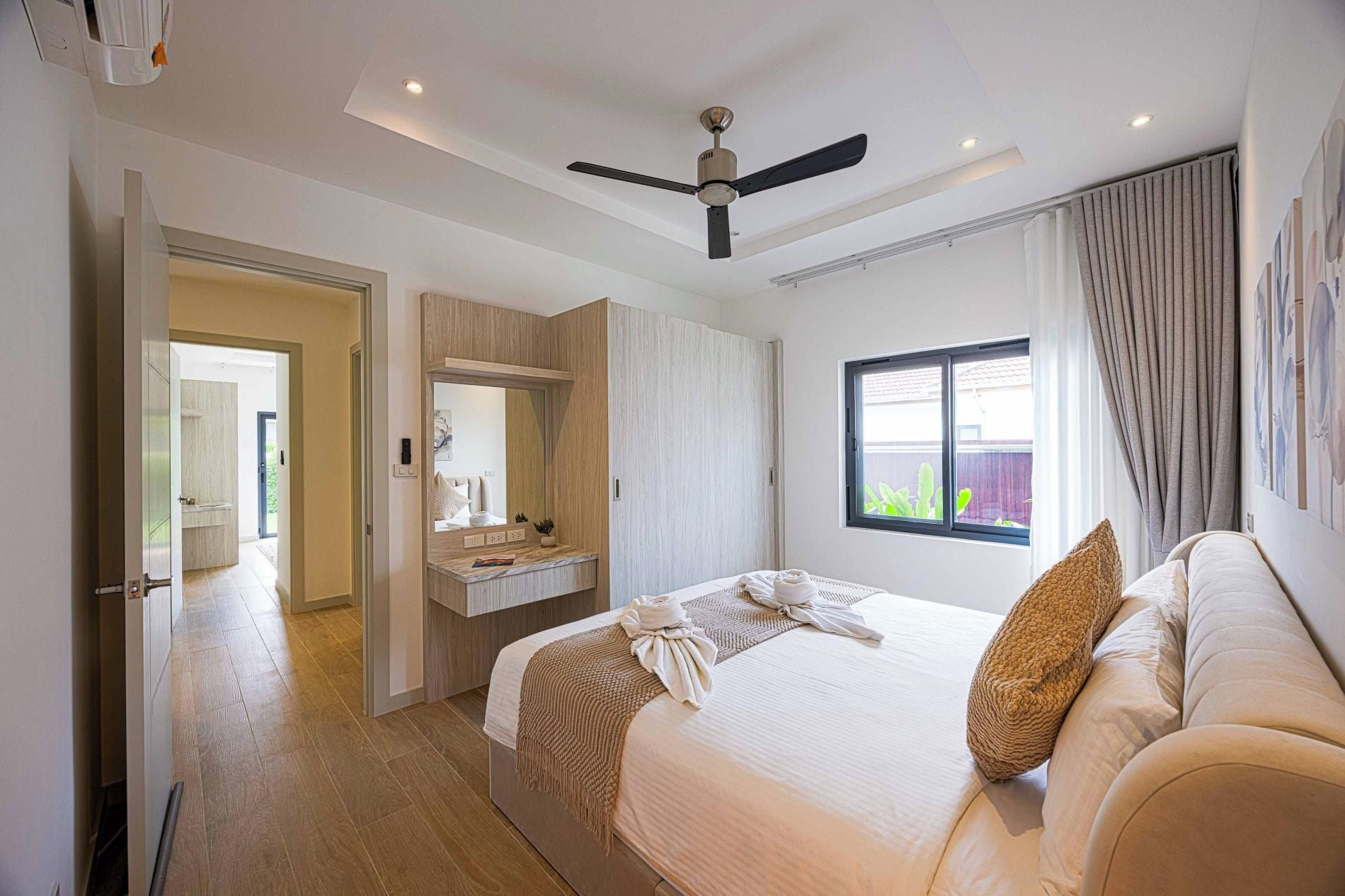 Une chambre d’hôtel avec un lit, une coiffeuse avec un miroir, un ventilateur de plafond et une fenêtre donnant sur un paysage verdoyant.