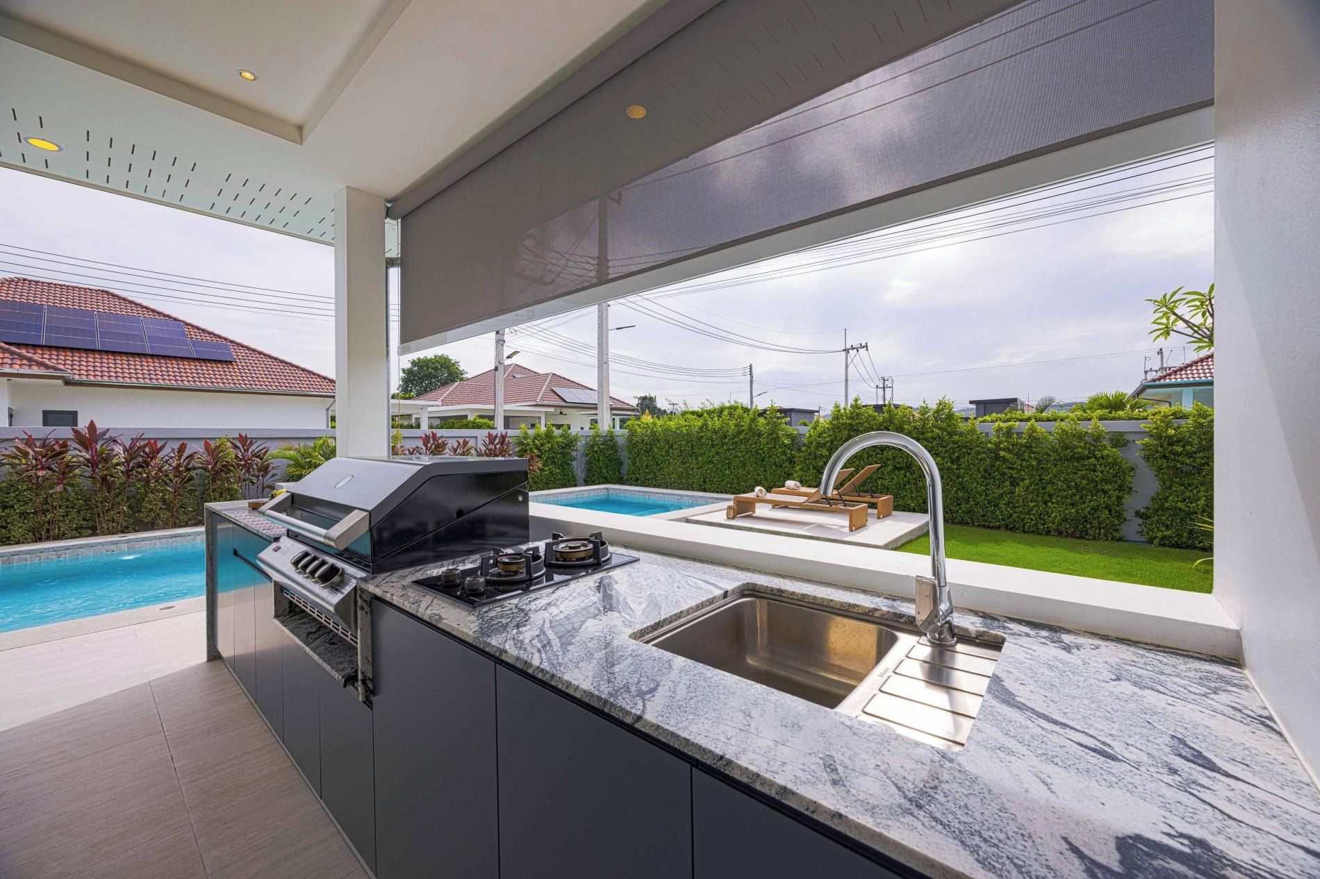 A modern kitchen with a built-in grill, a large window overlooking a swimming pool, and a view of neighboring houses.