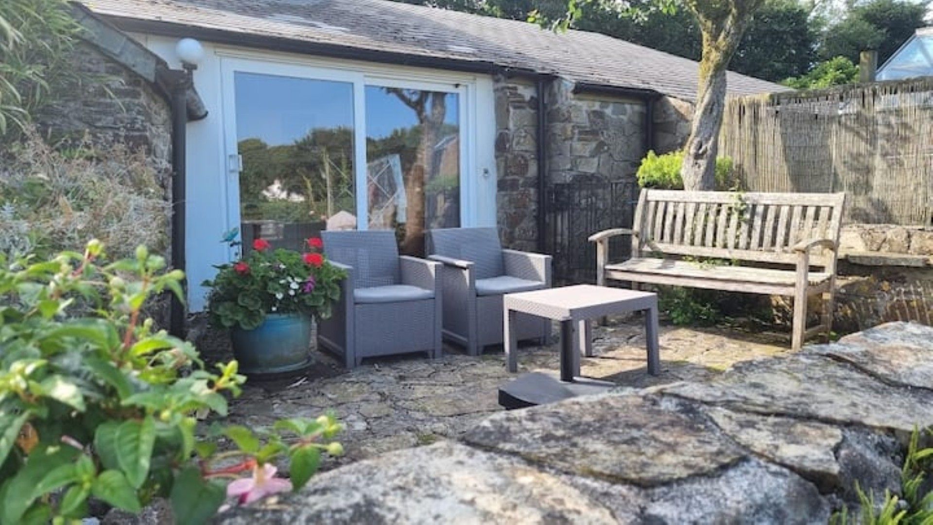 A patio with a stone table, two chairs, a wooden bench, and a potted plant with red flowers.