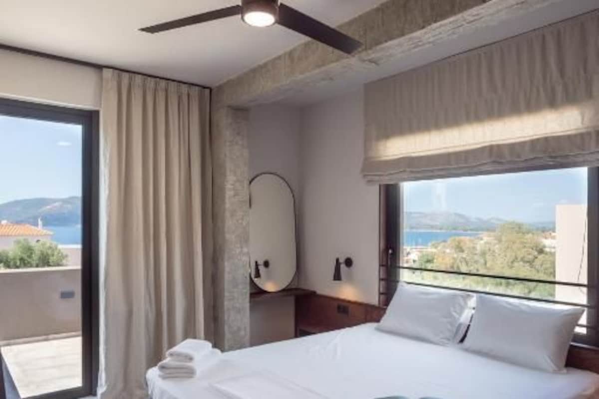 A modern hotel room with a large bed, a ceiling fan, a view of the city, and a balcony.